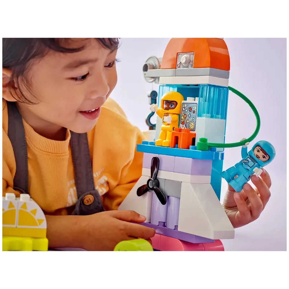 A smiling child in a yellow shirt interacts with a colorful LEGO® DUPLO® Space Shuttle playset, featuring two astronaut figures. The child holds a blue astronaut while another in orange stands inside the shuttle, surrounded by vibrant building blocks.