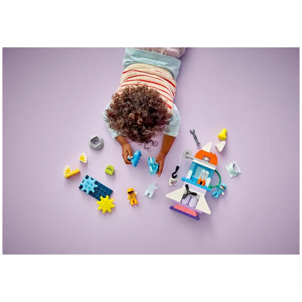 A child with curly hair kneels on a purple surface, focused on assembling colorful LEGO® DUPLO® pieces, including a rocket ship and accessories. Surrounding the child are various building blocks, astronaut figures, and playful shapes, inviting imaginative space adventures.