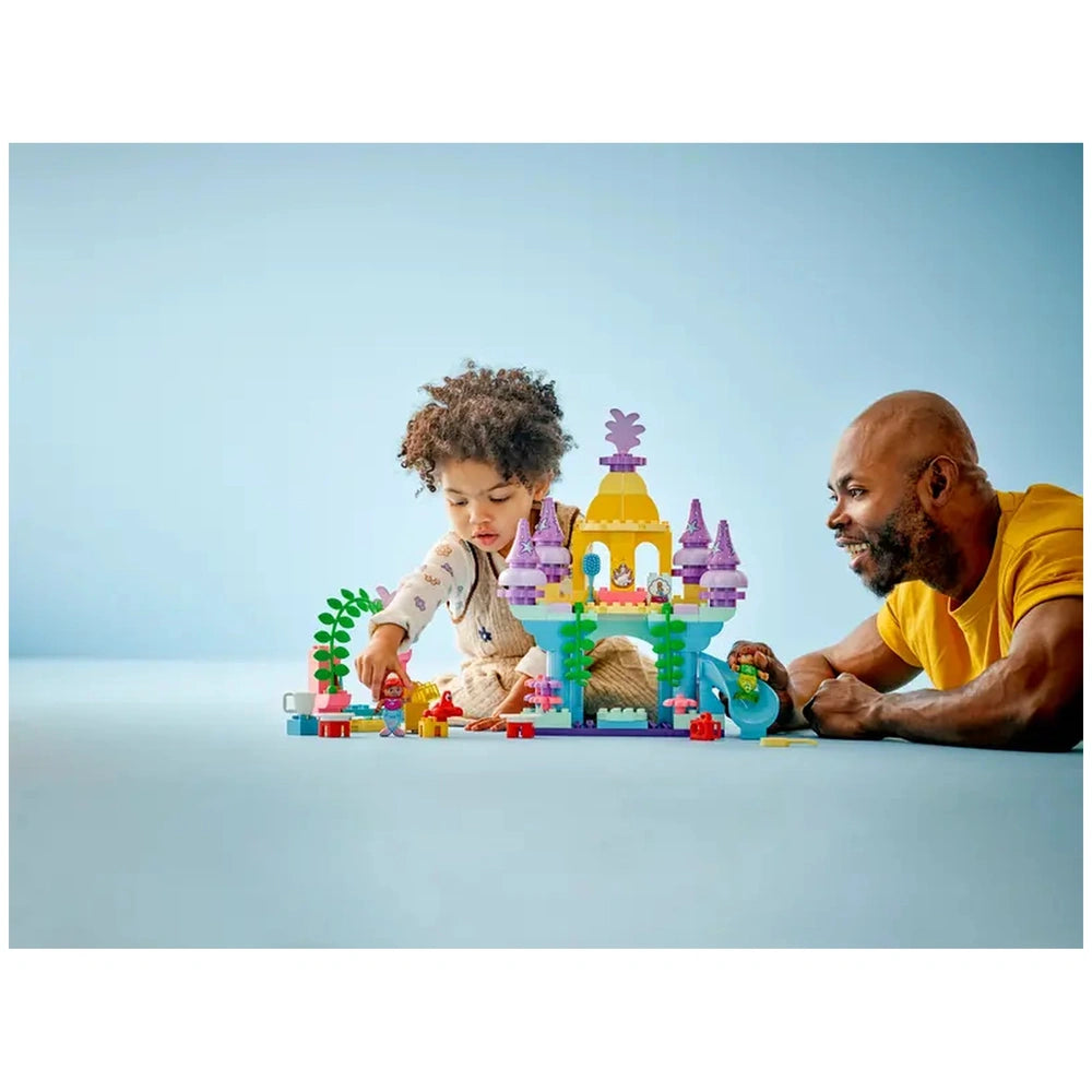 A child and a man play together with LEGO® DUPLO® Ariel's Magical Underwater Palace, surrounded by vibrant building blocks and figures.