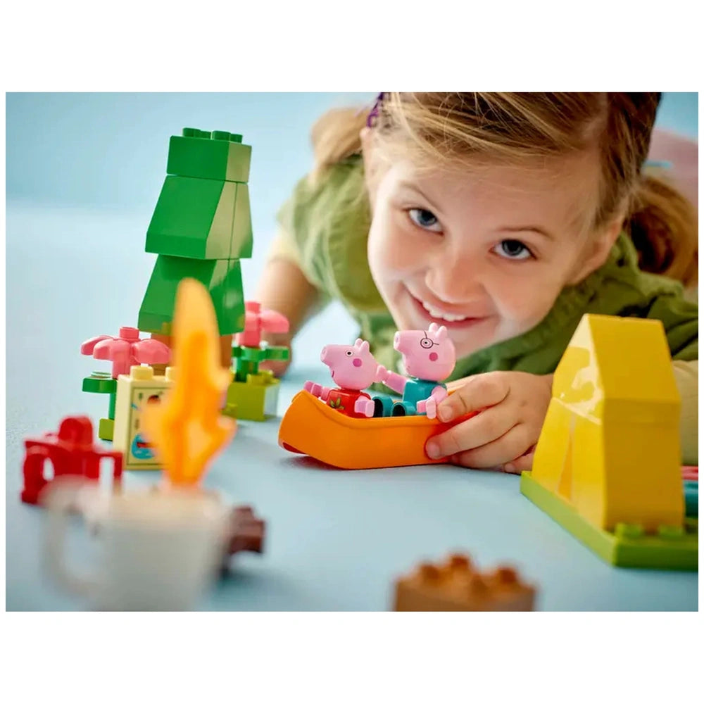 Young girl smiling while playing with the LEGO® DUPLO® Camping Trip set, featuring Peppa Pig and Daddy Pig in a canoe surrounded by colorful blocks and flowers.