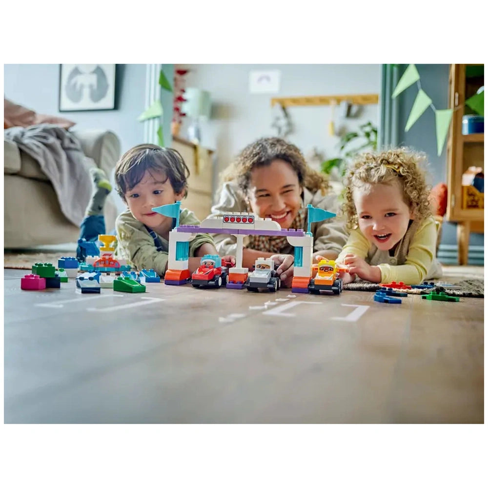 Three children play with Lego Duplo F1® Team Race Cars on the floor, surrounded by colorful blocks and a podium with flags.