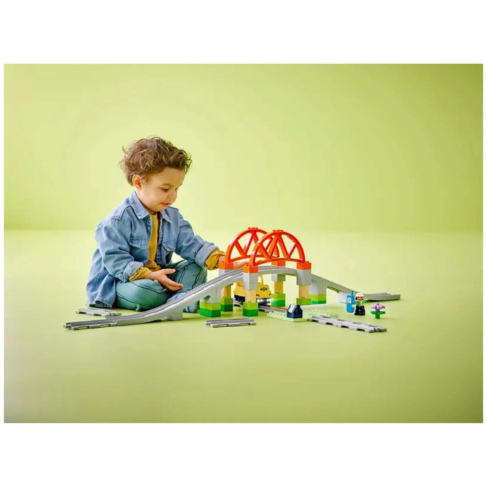 A young child in a denim jacket plays with the LEGO® DUPLO® Train Bridge and Tracks Expansion Set, featuring colorful blocks and a raised bridge, on a light green surface. The child is focused on assembling train tracks and figures, promoting creativity and imaginative play.