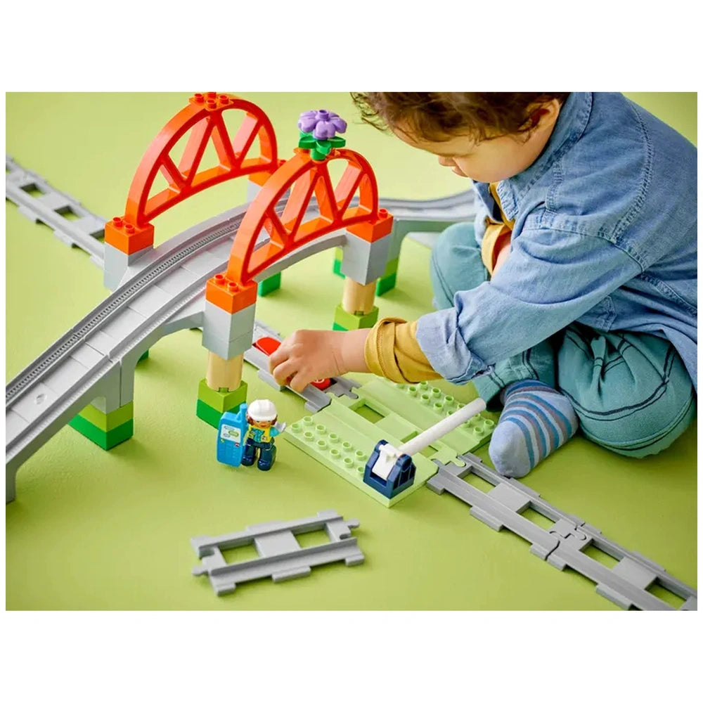 A child in a denim shirt plays with a colorful LEGO® DUPLO® Train Bridge and Tracks set, featuring a red arch bridge and a train barrier. The scene includes train tracks and small figures, set against a green background.