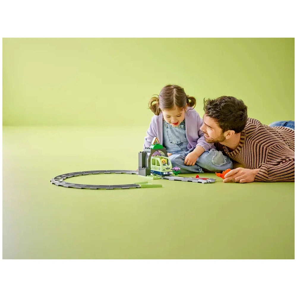 A child and an adult sit on a green floor, engaging with the LEGO® DUPLO® Train Tunnel and Tracks set. The child, wearing a purple sweater, smiles as they manipulate the colorful components, while the adult, in a striped sweater, leans in playfully. The set includes a tunnel and circular tracks, enhancing imaginative playtime.