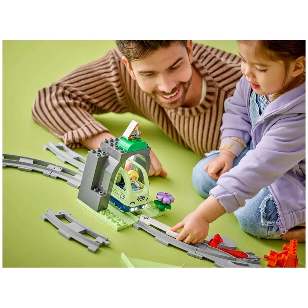 A smiling adult and a young girl play together on a green surface, interacting with the LEGO® DUPLO® Train Tunnel and Tracks Expansion Set. The set features a colorful tunnel and grey train tracks, along with a small flower and a warning sign. The girl points as they explore different track configurations, highlighting the imaginative and educational aspect of the toys.