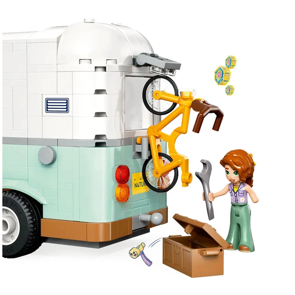 A LEGO camper van in pastel colors with a yellow bicycle attached to the back. A minidoll with curly brown hair holds a wrench while standing next to a brown toolbox. Bright accessories and details add to the playful scene, suggesting a fun camping adventure.