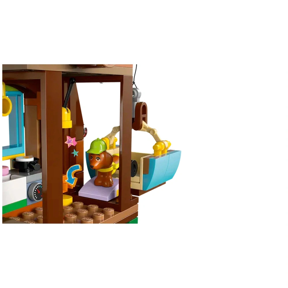 A playful brown dog wearing a green helmet sits on a purple cushion inside a LEGO tree house, with a blue bucket nearby and colorful decorations in the background.