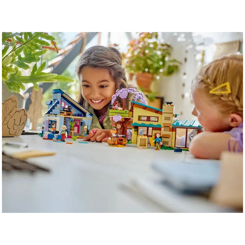 A girl with long hair smiles brightly while playing with LEGO® Friends Olly and Paisley's Family Houses, surrounded by colorful mini-dolls and detailed houses. Another girl leans on the table, observing the scene.