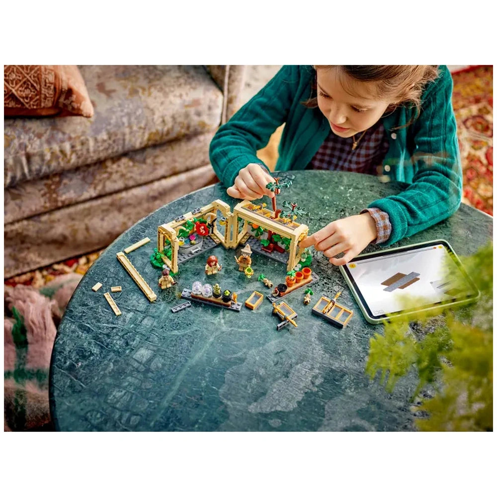 A child wearing a green sweater assembles the LEGO Hogwarts™ Herbology Class set on a round table, surrounded by colorful plant pieces, minifigures, and gardening tools. An instructional tablet is nearby, displaying building instructions.