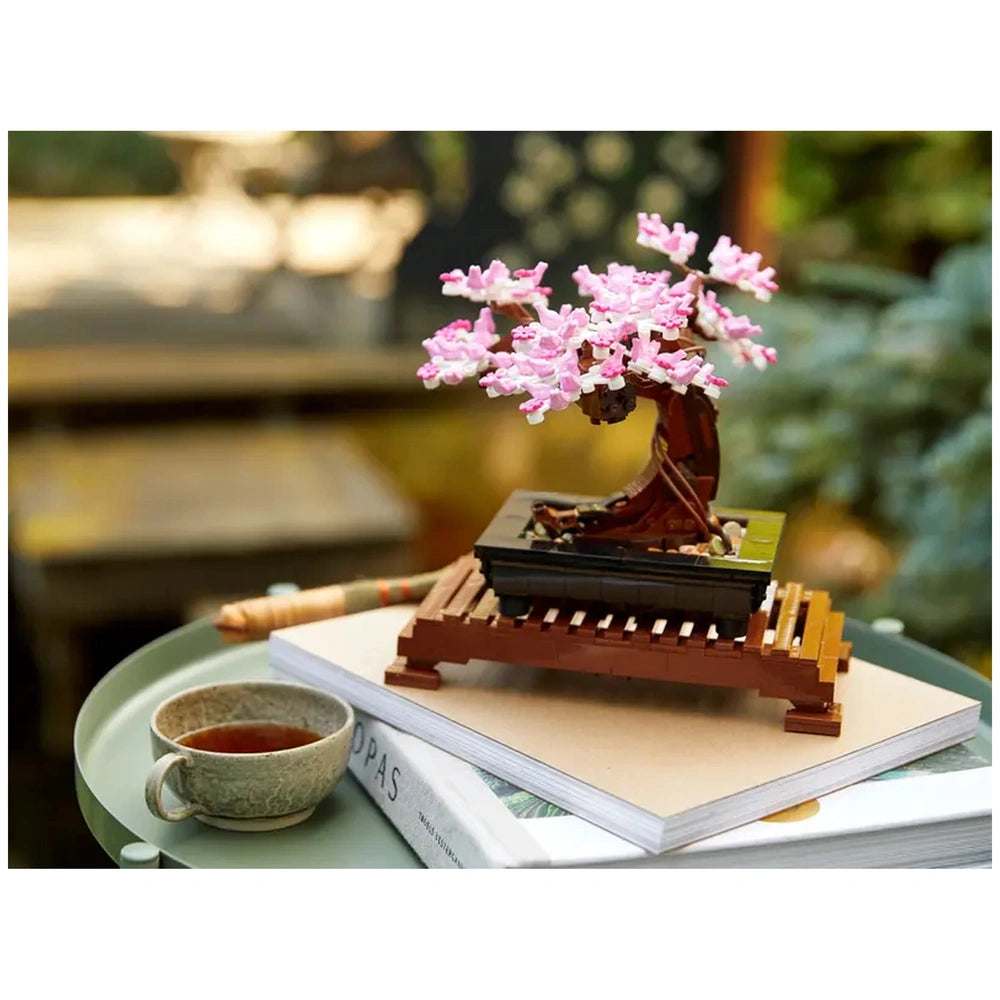 A detailed LEGO® Bonsai Tree model with pink blossoms sits atop a wooden stand. A small cup of tea rests beside it on a stack of books, surrounded by a tranquil garden setting.