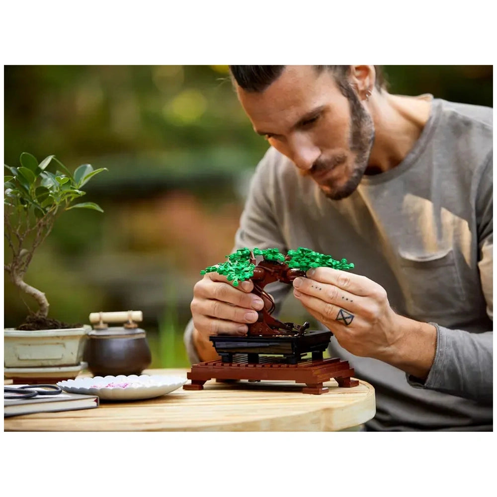 A person carefully assembles a LEGO® Bonsai Tree model, surrounded by a real bonsai plant and pottery, on a wooden table outdoors.