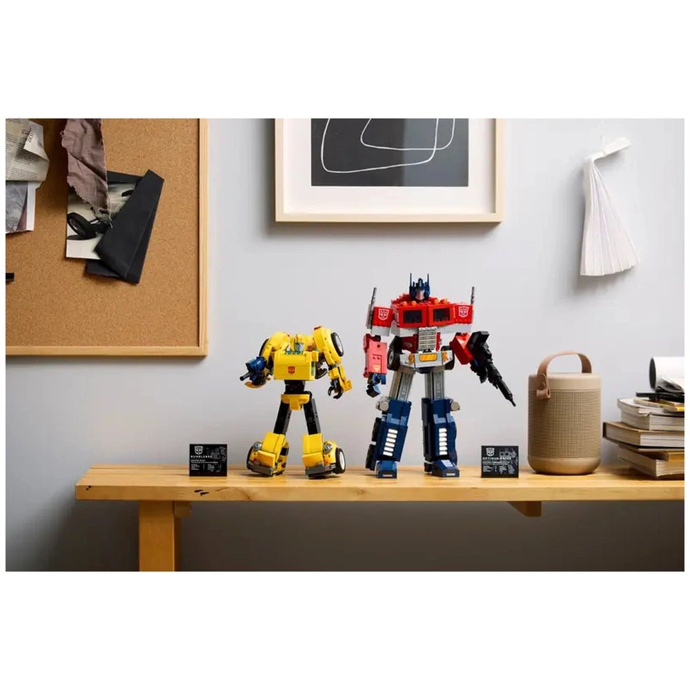 Bumblebee and Optimus Prime LEGO® Transformers figures stand on a wooden table, with a corkboard and a modern art print in the background.