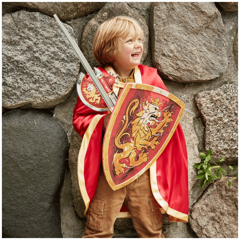 Child wearing a red Knight cape with a gold lion emblem, holding a matching shield, smiling in front of a rock wall.