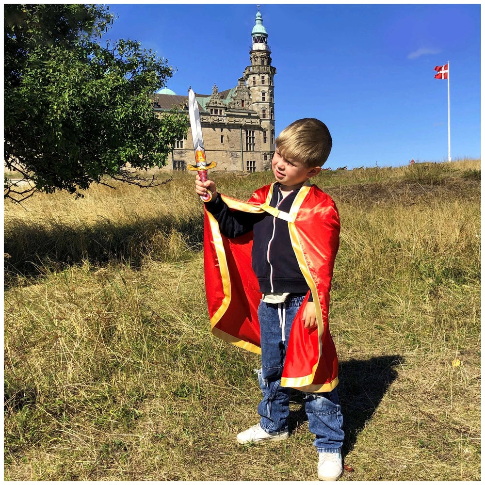 Young boy in a red cape with gold trim holds a Liontouch mini sword with a gold handle in a dry grass field.
