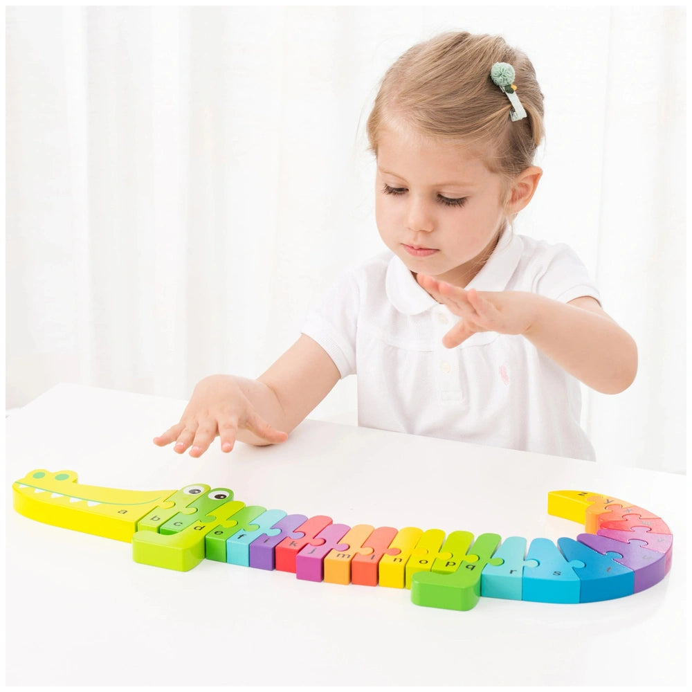 Alphabet puzzle by New Classic Toys on a white table, with a girl in a white shirt and green hair tie touching it.