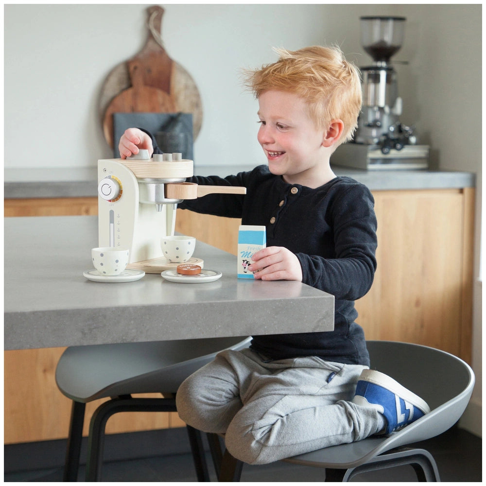 Child playing with a white New Classic Toys coffee machine at a wooden table surrounded by colorful plastic kitchen toys.