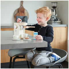 Child playing with a white New Classic Toys coffee machine at a wooden table surrounded by colorful plastic kitchen toys.