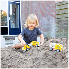 Child in blue t-shirt plays with yellow construction vehicle on sandy ground near a red brick building with white window fram