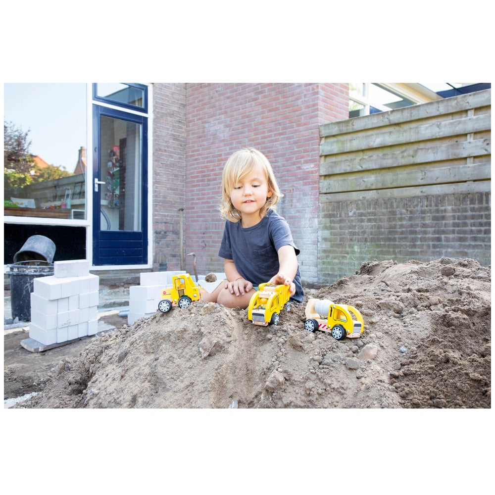 Construction Vehicles Set with a young child in a gray t-shirt playing with a yellow plastic toy truck on dirt near a brick b
