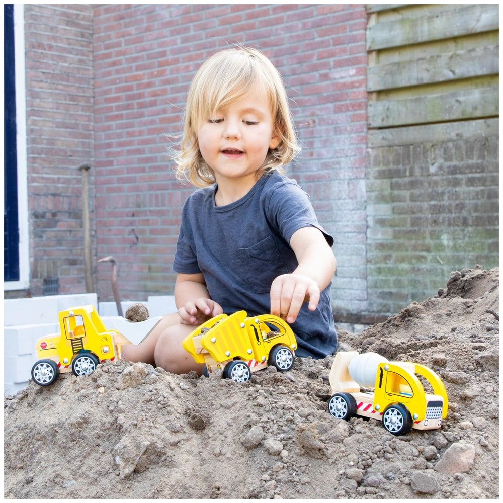 Child with fair skin and blonde hair in dark blue t-shirt plays with a yellow toy truck against a red brick wall.