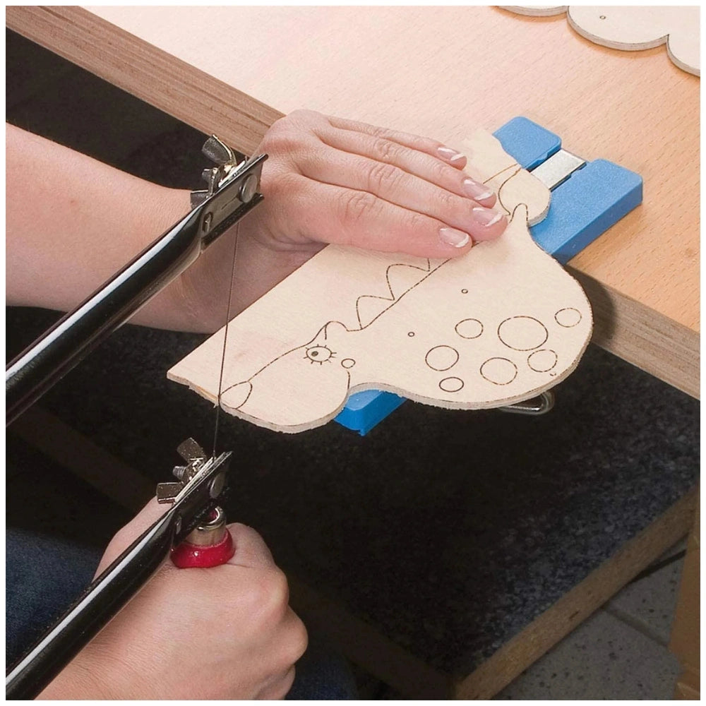 Hands using a fretwork saw to cut a dinosaur shape from wood, held on a blue clamp on a wooden table.