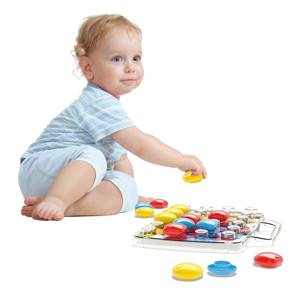 Quercetti FantaColor Baby activity toy with a young child in a light blue shirt holding a yellow object on a clear tray of co