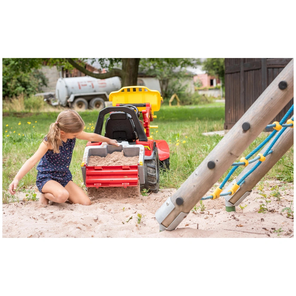Rolly Toys John Deere riding toy in a red sandbox with a girl playing, surrounded by green grass and a gray truck.