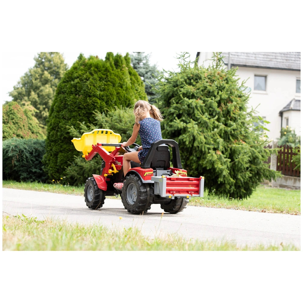 Child on a red Rolly Toys tractor with a yellow bucket, surrounded by greenery and a white house in the background.