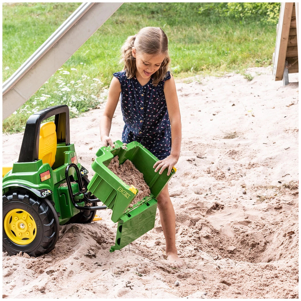 Girl in a blue floral dress playing near a tan sandbox on green grass with Rolly Toys John Deere riding toy nearby.