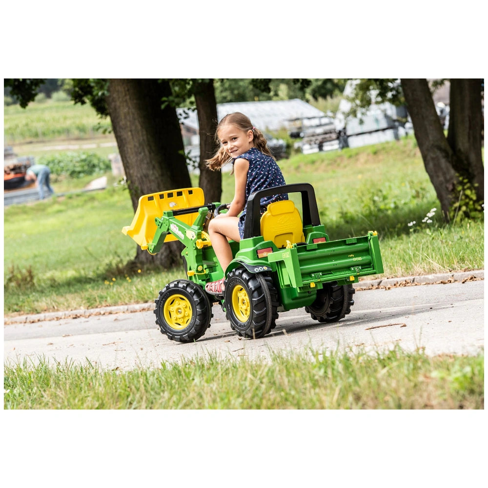Girl in blue shirt and pink shoes sits on a green and yellow Rolly Toys tractor on a grey road surrounded by trees.