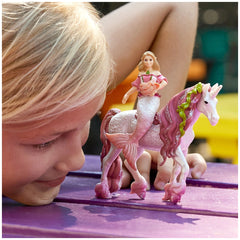 Mermaid Feya rides a unicorn on a purple surface, as a child with blonde hair leans over to admire the toy figures.