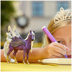 Schleich Star Pegasus Mare toy figure is featured alongside a young girl with a tiara, drawing with a purple pen at a yellow 