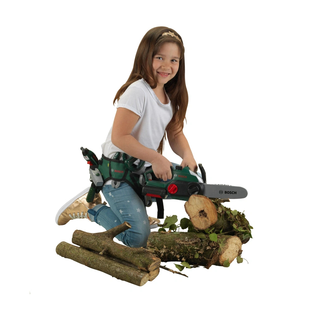 Theo Klein Bosch chain saw held by a smiling girl in a white t-shirt and blue jeans, standing on moss-covered logs.