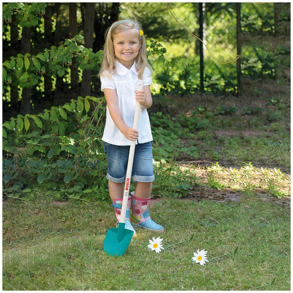 Girl in a white shirt and blue jeans holding a green shovel in a garden with white flowers and green grass.