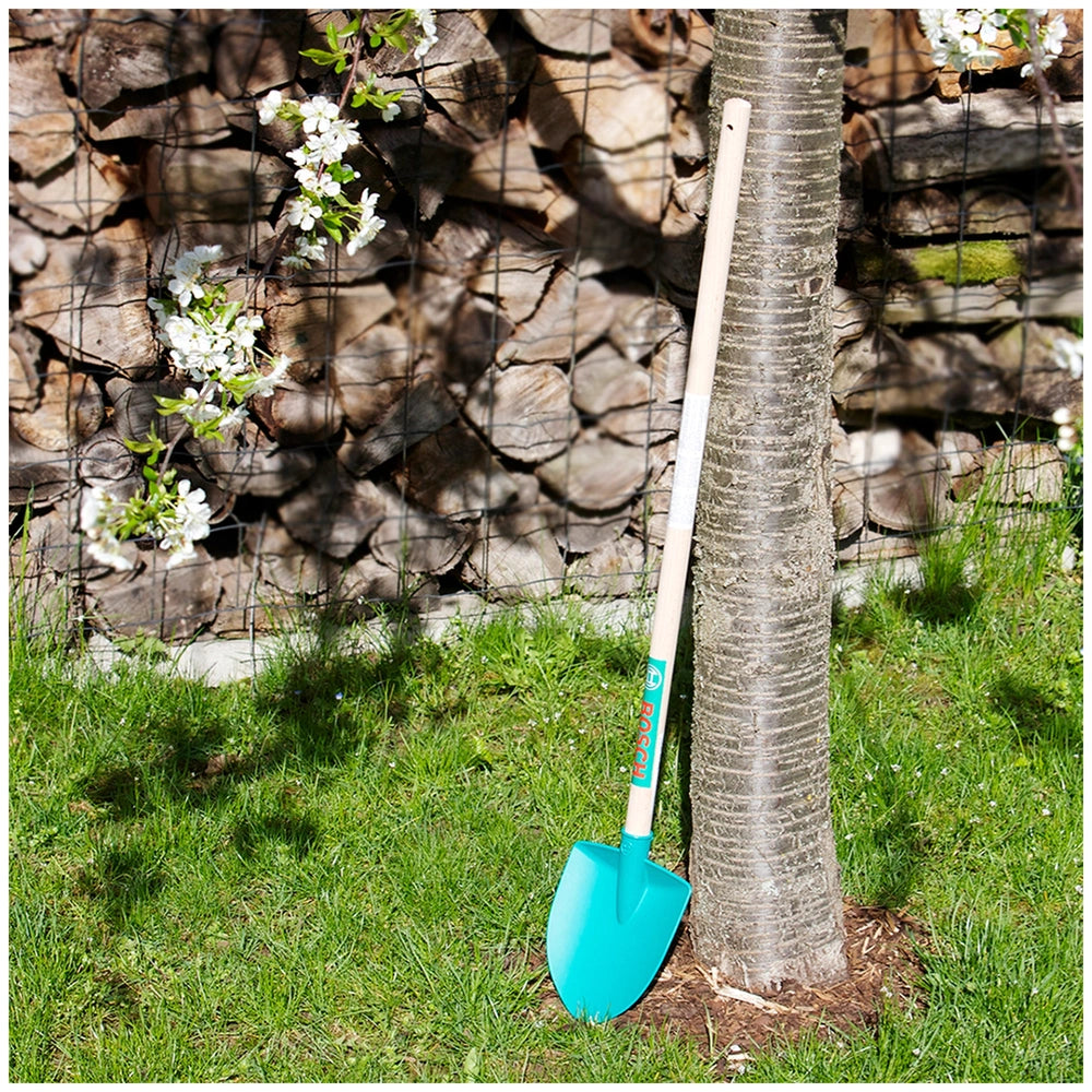 Blue Bosch garden shovel placed on green grass near a wooden fence, tree trunk, and white flowers in a vibrant outdoor settin