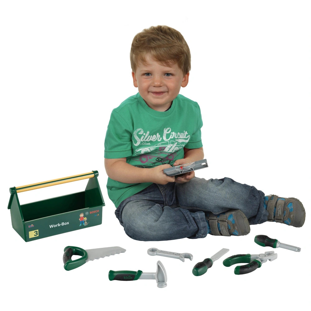Theo Klein Bosch Work Box with child holding gray scissors, sitting on white floor next to green box with yellow handle.