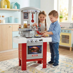 Theo Klein Miele kitchen set with silver and red oven, white countertops, and two children role-playing in a light blue theme